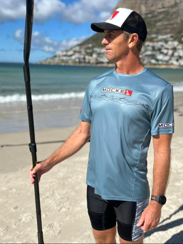 Man on a beach wearing a blue athletic shirt and black cap, holding a paddle.